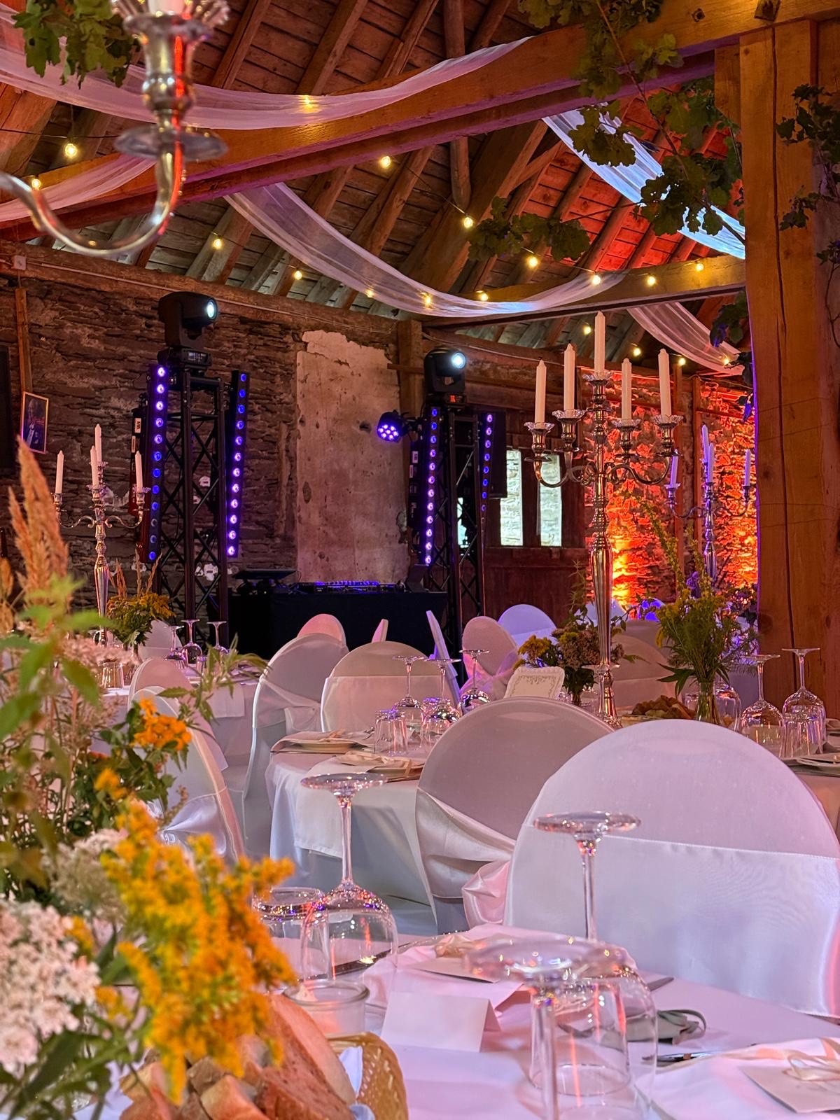 Festlich gedeckte Hochzeitstafel in einer Scheune, dahinter DJ-Pult mit Traversen und Moving Heads